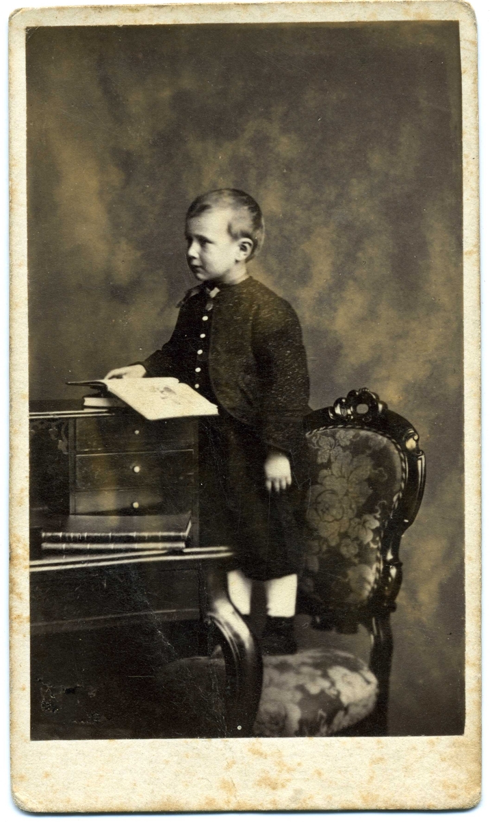 Kabinettsfotografi: Erik Dahlström, en liten pojke, står på en stol med uppslagen bok ovanpå ett skrivbord.