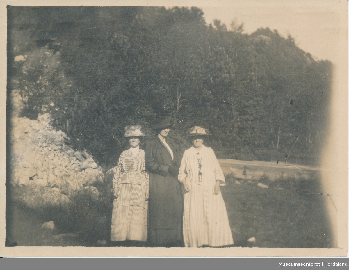 amatørfoto av tre kvinner i kle frå tidleg 1900-tal med hatar, to i ljose kle og ei (i midten) med mørke kle. Dei står på ein mark framfor skog og med eit vatn i bakgrunnen