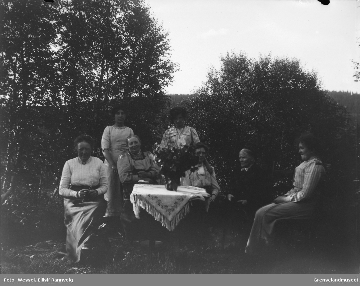 En gruppe kvinner fotografert ved et bord ute i en hage, ukjent sted. En kvinne sitter med strikketøy.