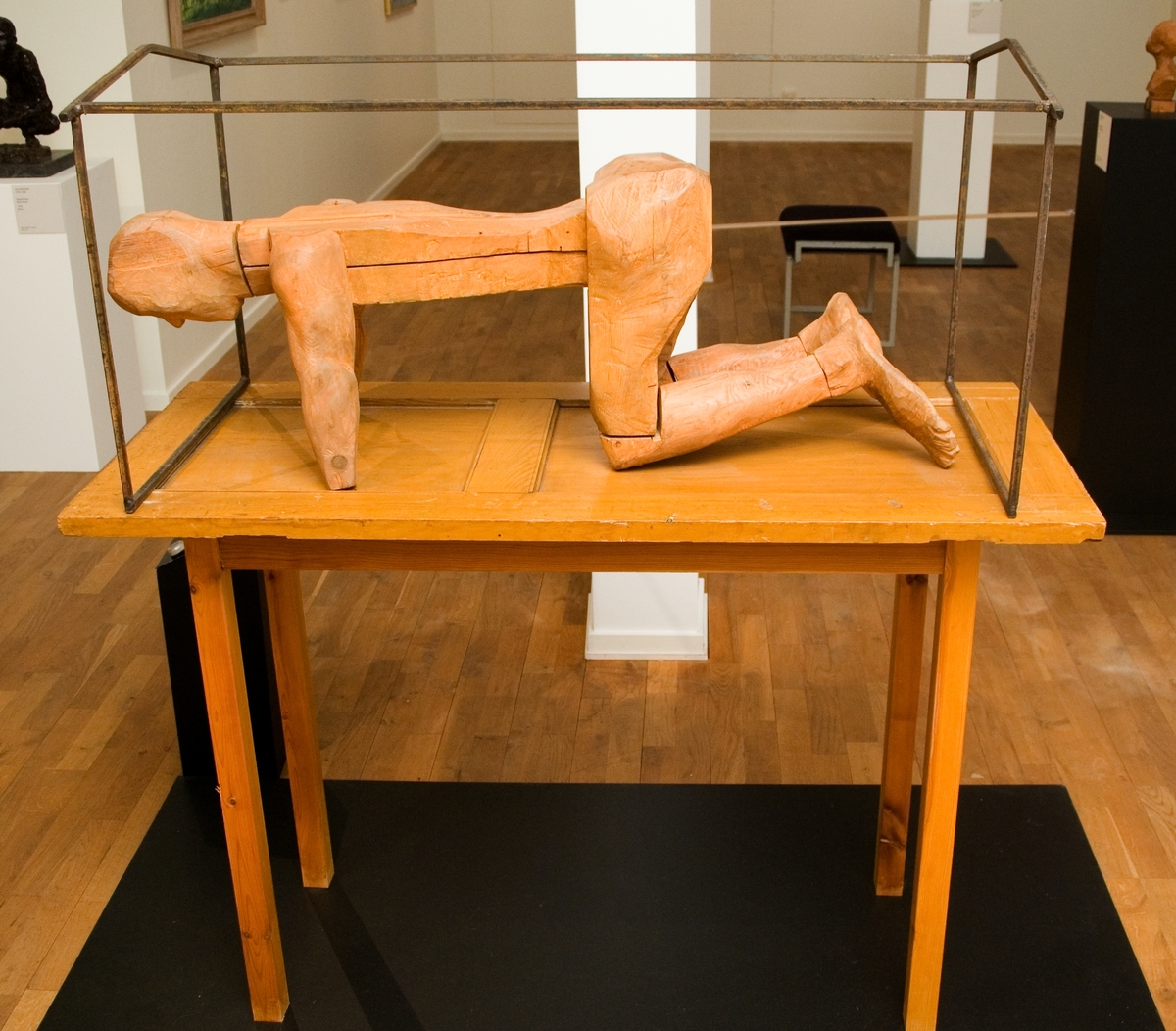 Träskulptur av Torsten Renqvist "Mannen i buren", 1968. Sockeln till skulpturen är uppbyggd som ett bord. Bordsskivan är en dörr som bärs upp av  fyra ben. På "bordet" en skulpterad mansfigur av trä, knästående. Han omges av ett järnstativ som symboliserar buren.
Skulpturen märkt på bordets undersida och mansfiguens bröst.