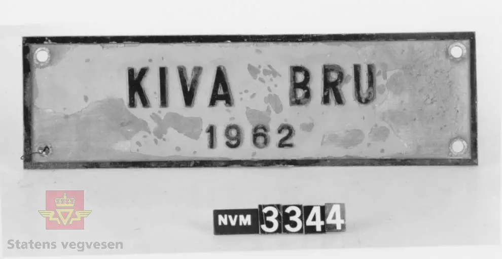 Rektangulært navnskilt med teksten : "KIVA BRU 1962 ". Lagd av støpt aluminum med svart tekst og bord på gul bunn. Skiltet har hull i hvert hjørne for innfesting. 