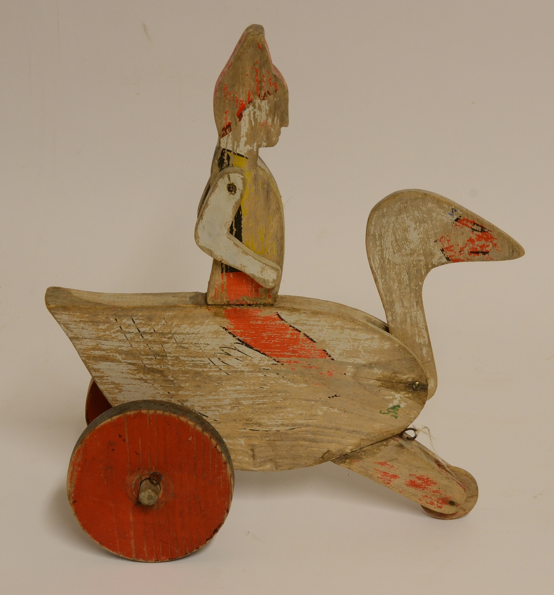 Leksak av trä i form av en gås med en pojke (Nils Holgersson) i röd luva på ryggen. Leksaken dras med ett snöre, rullar på tre trähjul. Målad i vitt, rött, gult, svart och brunt. Pojkens armar och gåsens huvud är svängbara samt fästa med spik.