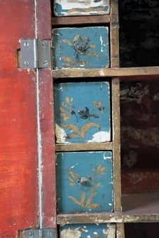 Hängskåp av trä med brutet krön och dubbellucka, blå grundmålning samt ornering i guld, svart och grönt. Dörrarna har landskapsmotiv inom målad rokokoinramning. Två hyllor i mitten med sex små utdragslådor på vardera sidan.