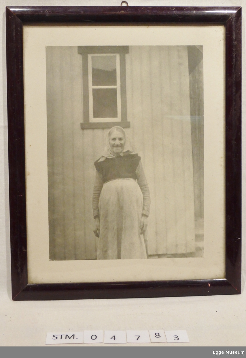 En gammel kvinne står foran et trehus. Hun har på seg kjole med forkle og skaut. Både kroppen og blikket er rettet mot kameraet. På veggen bak henne er det en vindu med to ruter. Til høyre i bildet kan det skimtes trær.