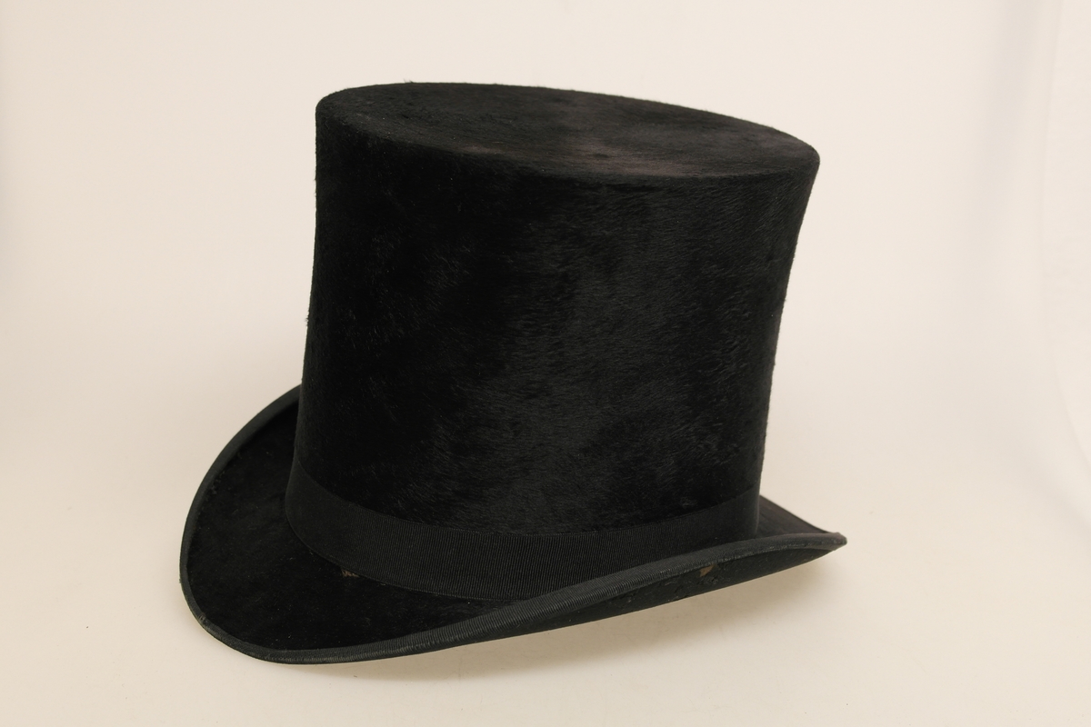 Svart flosshatt i en type fløyel (kastor/beverhår?). Hatten har svart ripsbånd med svart, rektangulær spenne festet på den ene siden. Innvendig er hatten kledd med papir og et silkeaktig stoff i purpur. I bunnen er det et gull-/kobberfarget stempel.