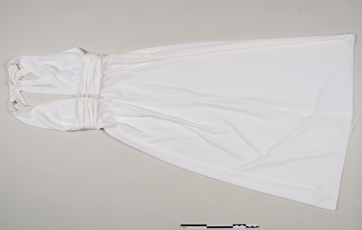 Kremhvit kjole sydd av polyesterstoff. Det er en glidelåsåpning bak og 5 pynteknapper foran. Kjolen har en V-utringning og ingen ermer.