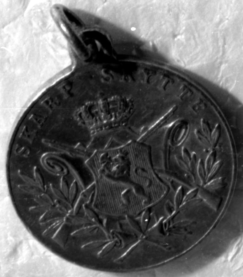 Rund medalje med det norske riksvåpenet med krone i midten over to gevær i kryss, og laurbærblad rundt.