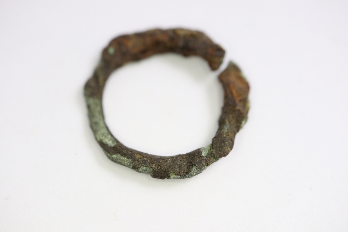 Fingerring av brons som är ornerad med graverade snedstreck (repslagsimitation?). Den övre delen av ringen är något högre än den undre. Förorenad med rost och små trärester.