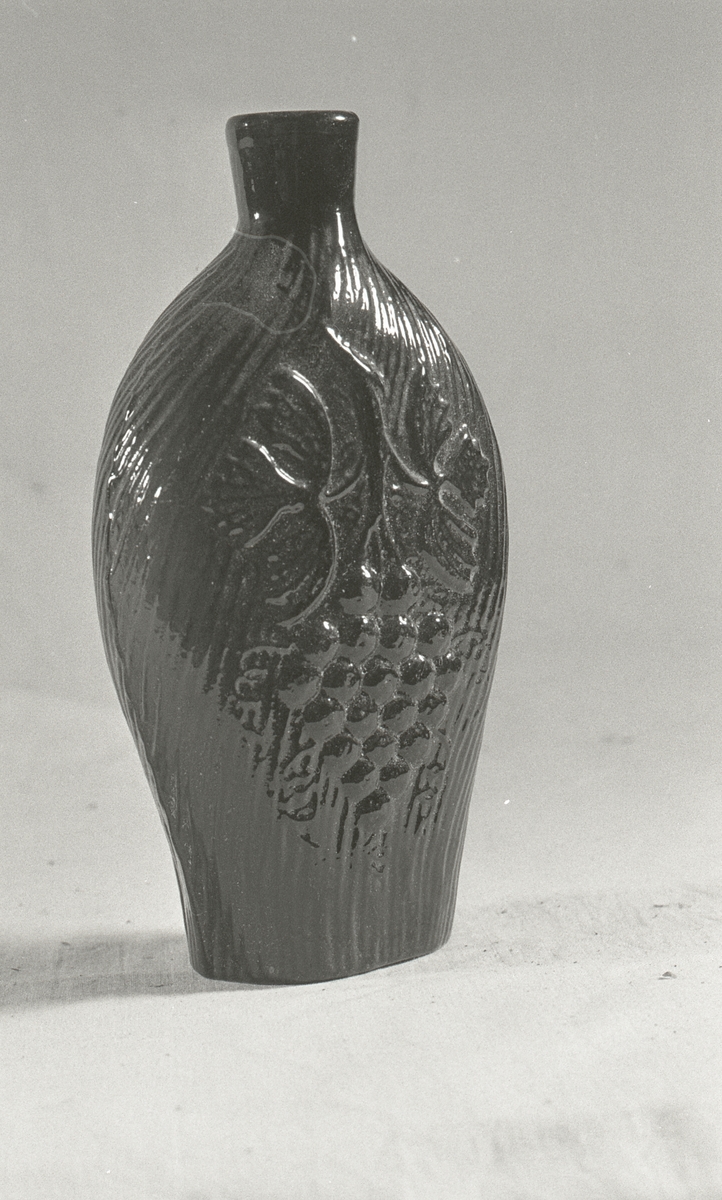 Glass, fiolett, riller, løve på ene sida og drueklase på den andre. Støperand.

Brennevinsflaske som vanligvis kalles "løveflaske". Laget på et av de norske glassverkene, annen halvdel av 1800-tallet.