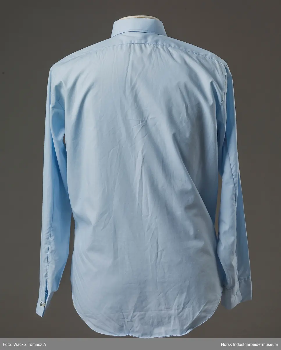 Lys blå, langermet uniformsskjorte i tekstil av 65% polyester og 35% bomull. Nedfelt krage. Lukking i front med 7 grå knapper. Brystlommer med klaff som lukkes med en grå knapp. Mansjett har et knappehull og lukkes med grå knapp.