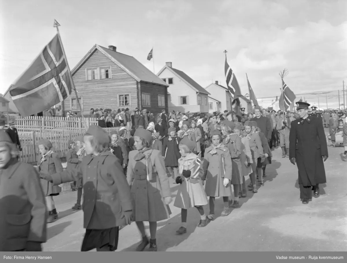 Vadsø 17 mai 1952. 17.mai-toget går gjennom Oscarsgate fra øst mot vest. Ser åssiden Melkevarden i bakgrunnen. Ser speiderjenter og barn med flagg. To politimenn i uniform.