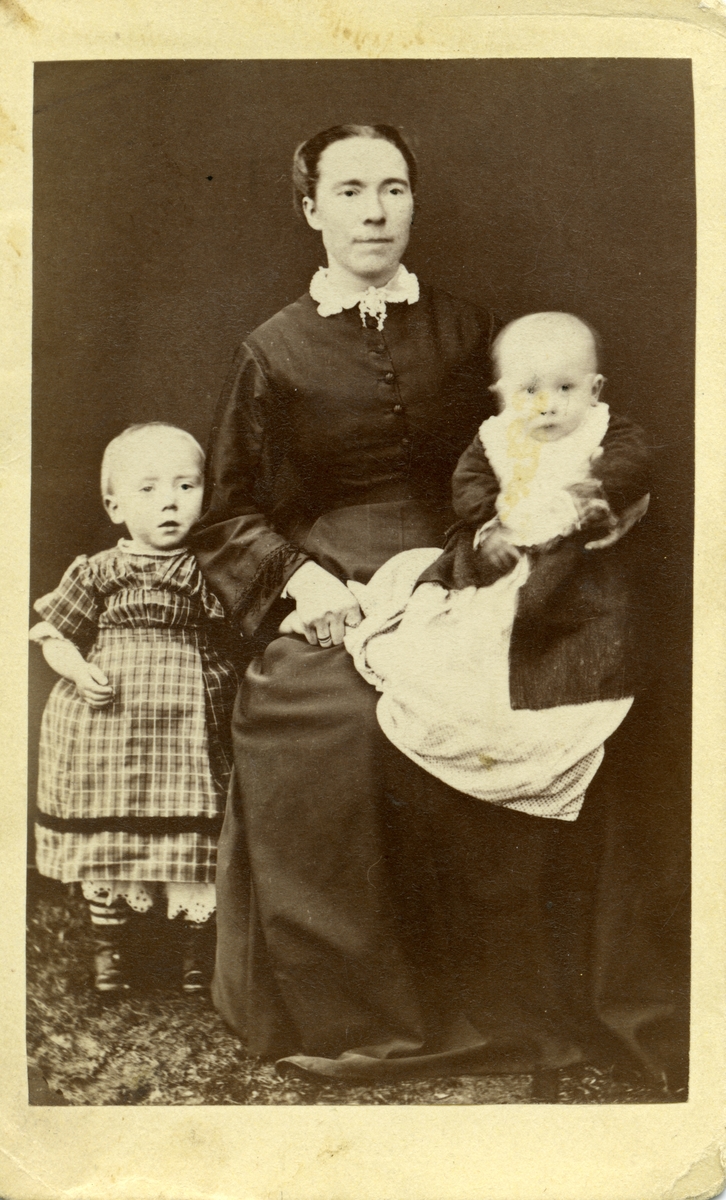 Portrett i helfigur av ei kvinne, ett spedbarn som sitter på fanget hennes og ei lita jente som står ved siden av.