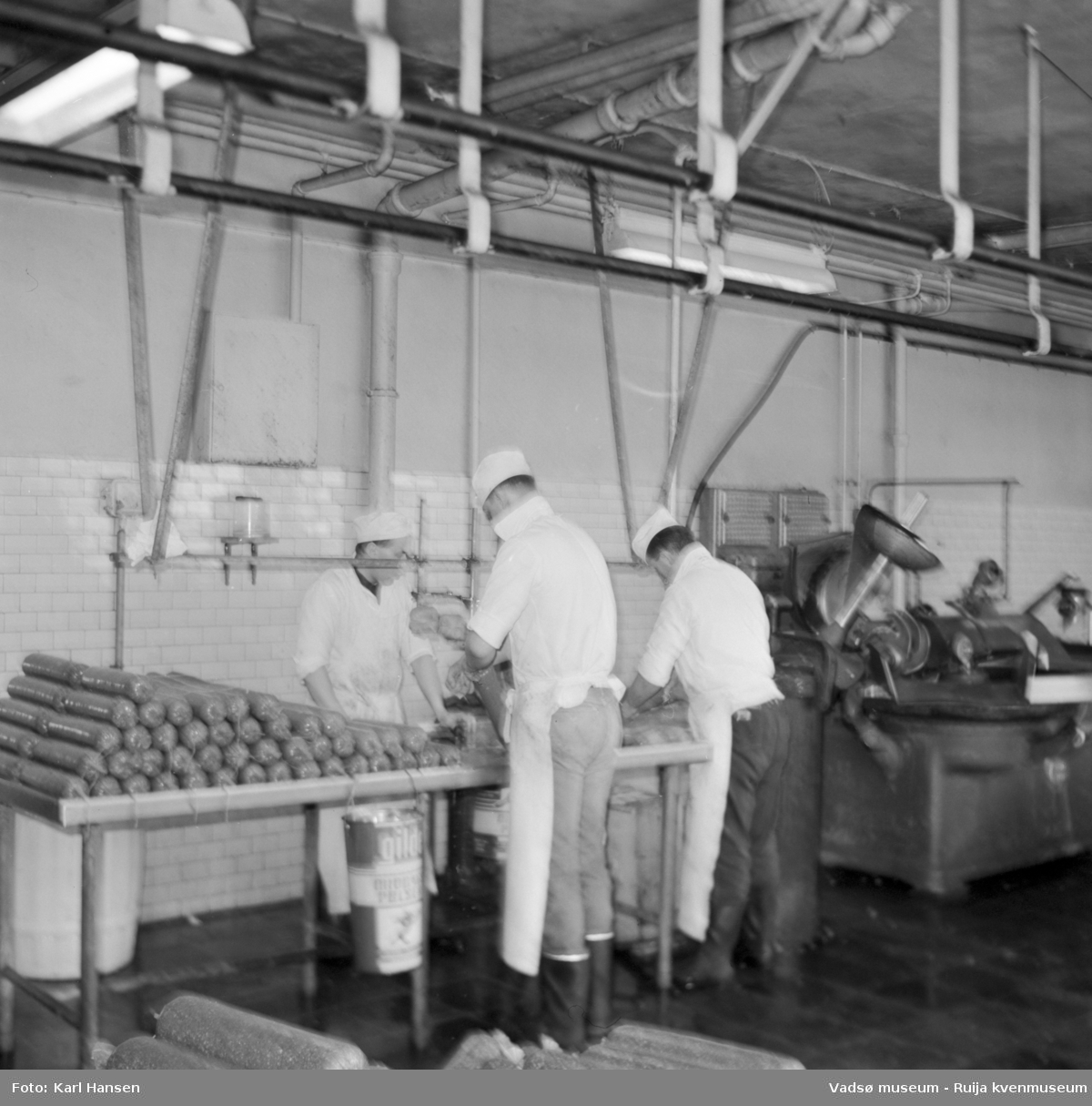 Nord-Norges salgslag Vadsø, 1964. Tre menn arbeider med spekepølseproduksjon i produksjonshallen. Fra venstre; Valter Kestilæ, Roy Aronsen og Ronny Johnsen.