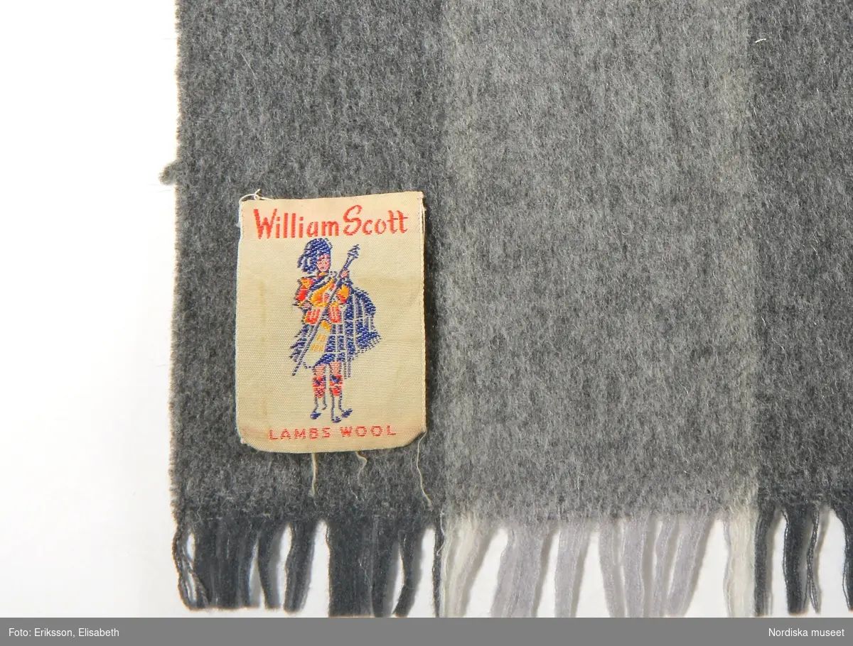 Halsduk, 1962-1968, av rutig, varp av vitt och två nyanser grått ullgarn. Inslag av grått, grönt, vitt och svart ullgarn, kort frans, text på påsydd etikett 'Wiliam Scott Lambs Wool'.