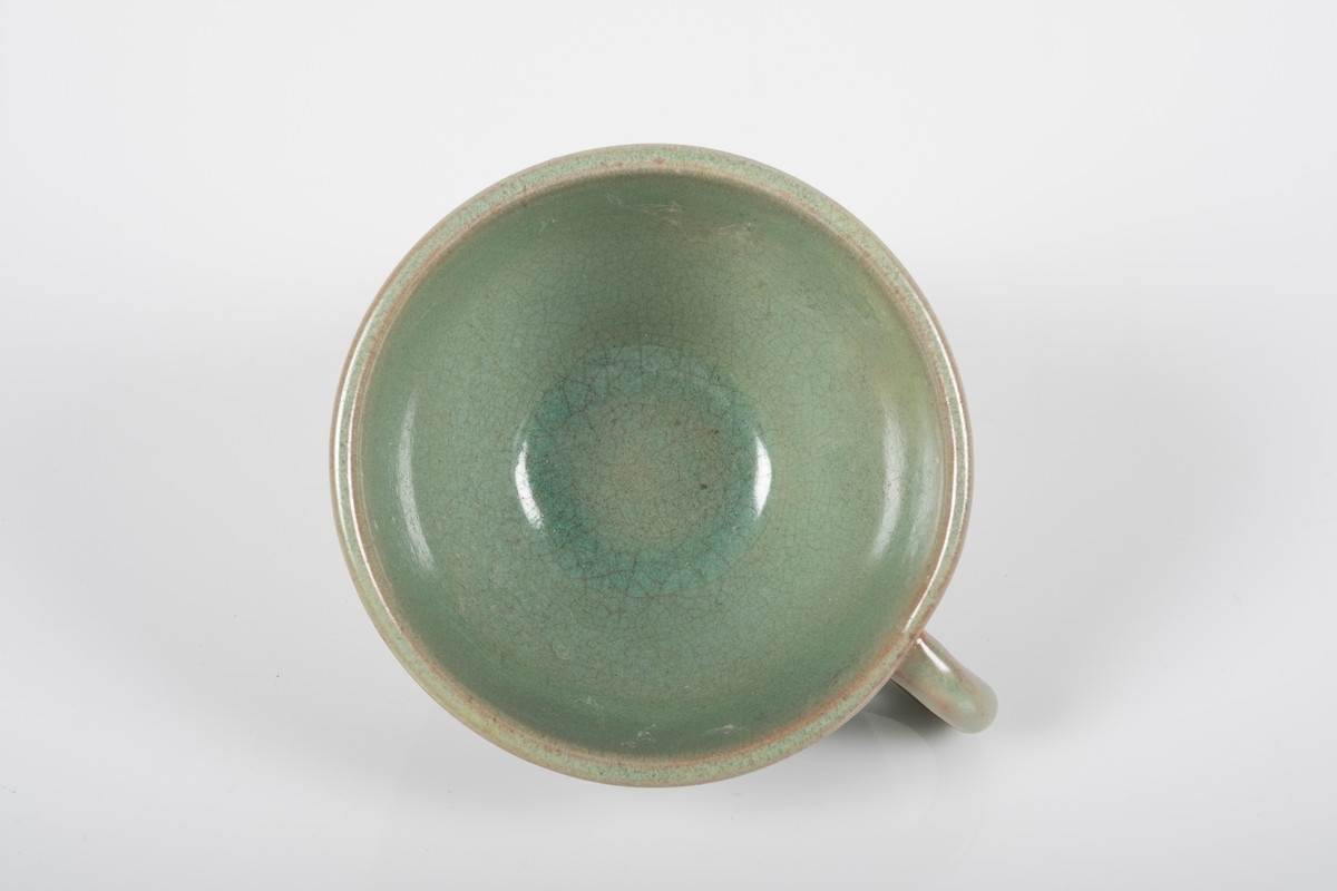 Rund kopp i keramikk med grønn lasur. Buet hank på koppen. Tre små knotter på undersiden av koppen, usikker funksjon.