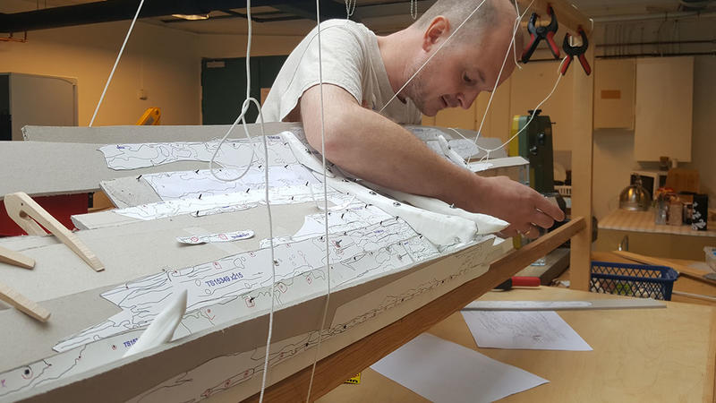 Arkeolog bøyer seg over og arbeider på innsiden av en uferdig båtmodell i plast og papp. Modellen er støttet opp i en treramme ved hjelp av klemmer og klyper i tre og plast, samt snører på flere steder. (Foto/Photo)