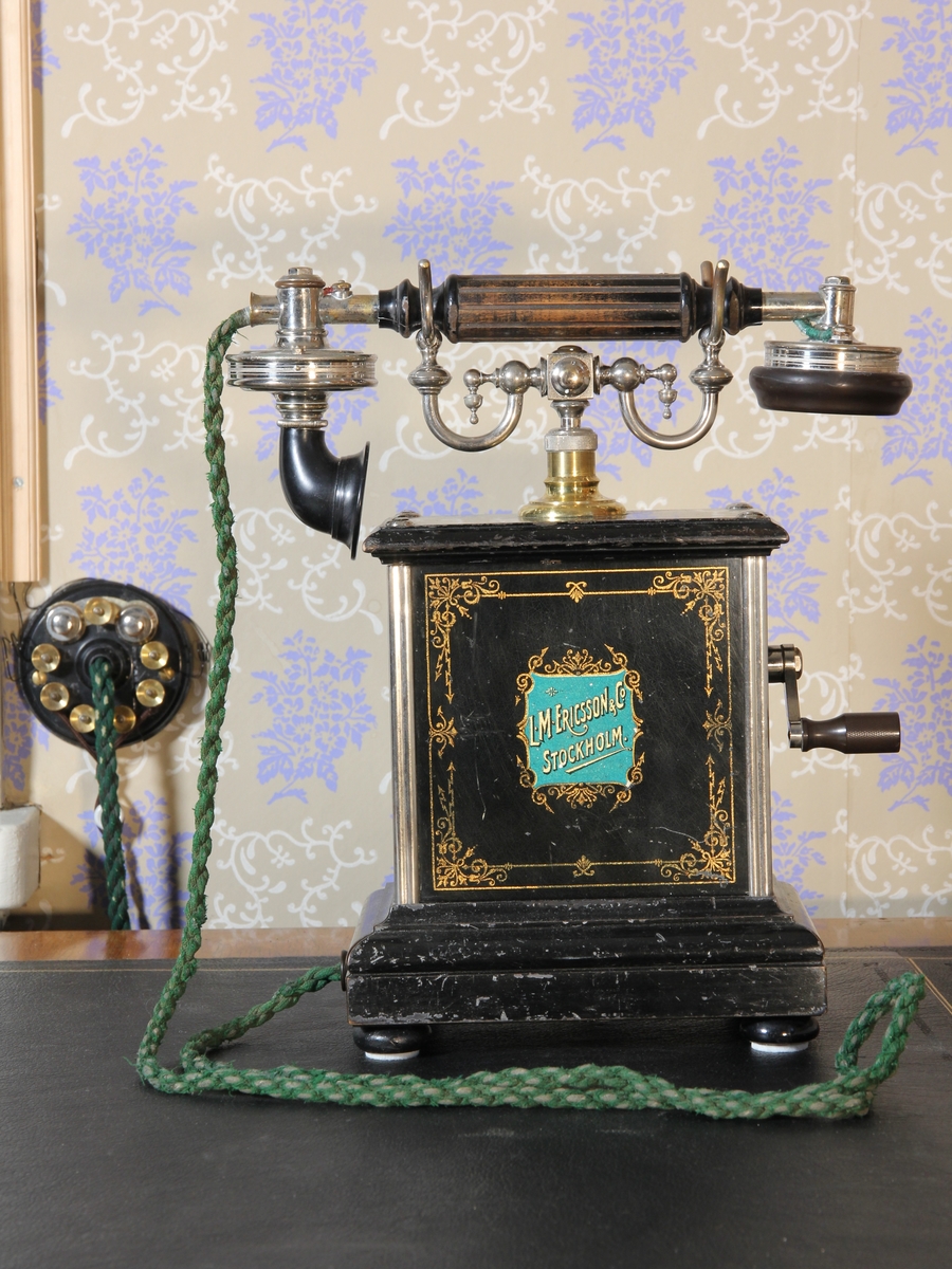 Telefon, bordsapparat med lur. Modell A 381 Jubileumsapparat (framtagen till L M Ericssons 25-årsjubileum 1901). Stående rektangulär med klyka av vitmetall. Svartmålad med dekor och text bland annat i guldfärg. Vev. Väggplint och grön sladd.