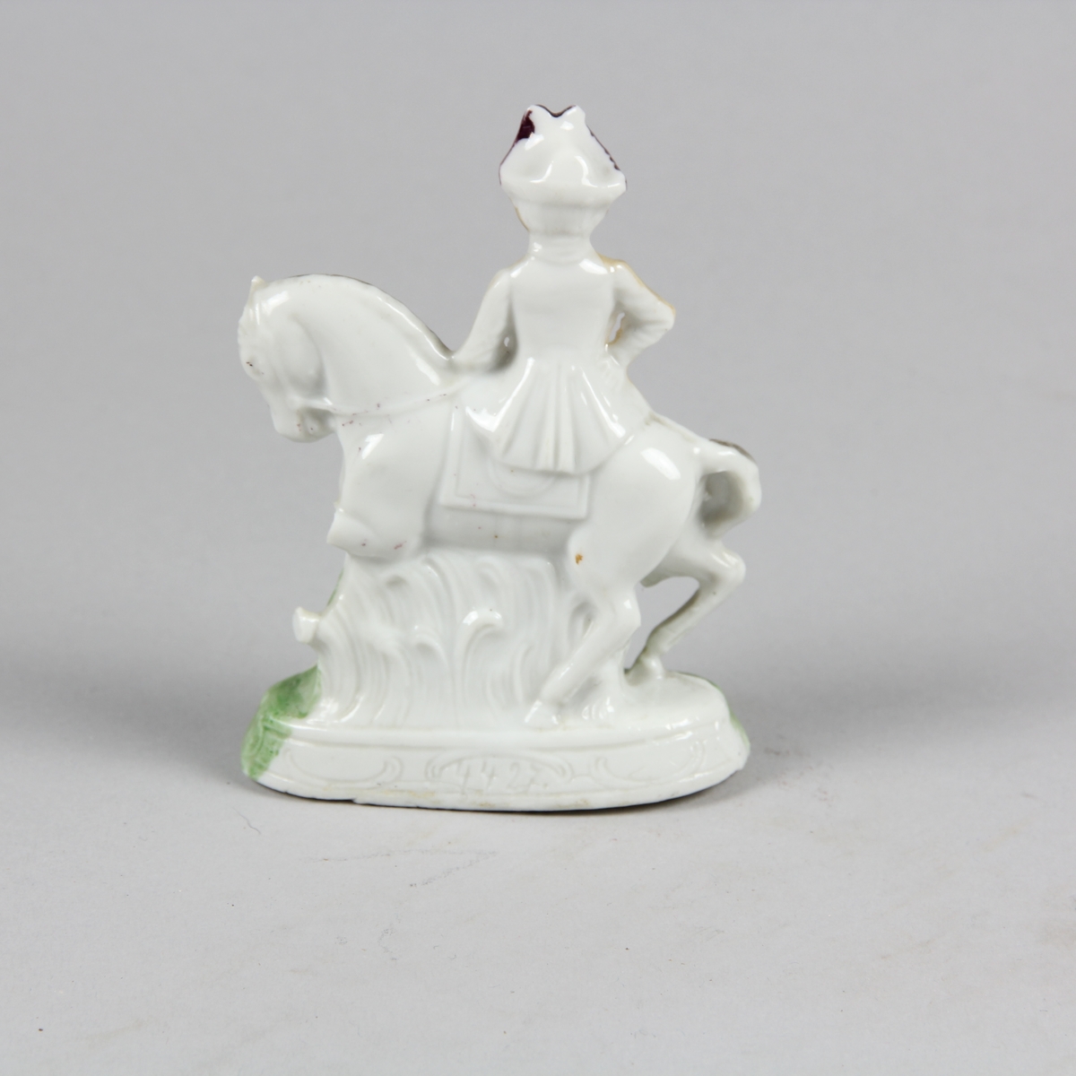Prydnadsföremål av vitt porslin i form av liten skulptur föreställande en kvinna som sitter på en häst i damsadel. Kvinnan är klädd i lång gulvit klädnad och lila hatt. Hästen vit med svart man. Grönt gräs och grön sockel. Endast framsidan, vänd mot betraktaren är bemålad.
