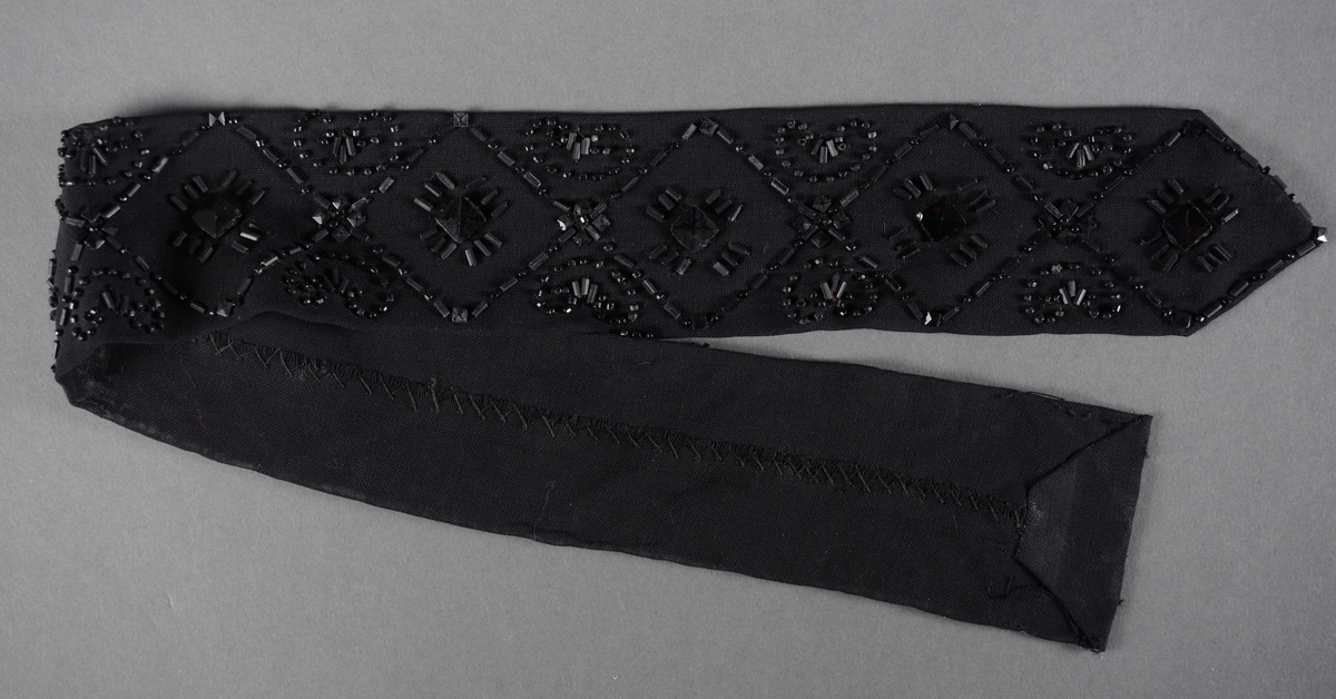 Drakttilbehør, muligens et belte, laget av svart chiffon og svarte glassperler. Det er håndsydd av dobbelt lag chiffon. Til perlebroderiet er det brukt firkantede, avlange og runde perler. Mønsteret er geometriske figurer.