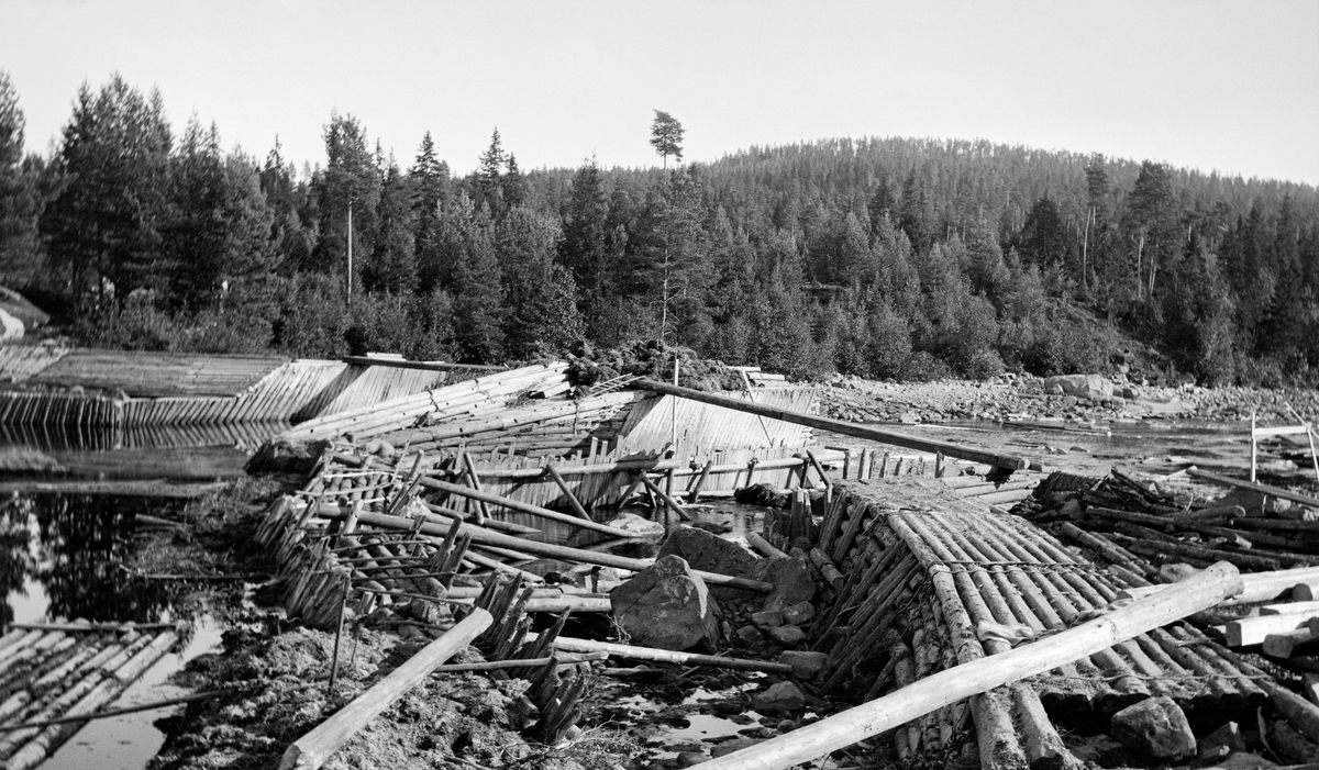 Første fase i ombygginga av Kjellåsdammen i Flisavassdraget i Åsnes i Solør (Hedmark) i 1915.  Fotografiet er tatt seinhøstes, på et tidspunkt da vannføringa i Flisa var meget lav, slik at konstruksjonene var lett tilgjengelige for anleggsarbeiderne.  Da dette fotografiet ble tatt var de «gamle» konstruksjonene - en steinfylt tømmerkistedam - under demontering.  Arbeidet foregikk i ly av en såkalt fangdam, som skulle holde vannmassene unna området der arbeidet foregikk.  Kjellåsdammen var en såkalt atthaldsdam som kunne demme opp den nedre delen av Flisavassdraget i en lengde på opptil fem kilometer.  Den dammen det avbildete anlegget avløste var bemannet med to damvoktere så lenge det pågikk fløting i Flisa.  Tidlig på 1900-tallet var man i tvil om den tilsynelatende begrensete nytteverdien Kjellåsdammen og bemanninga der hadde forsvarte driftsutgiftene.  Det ble snakket om at anlegget samlet lite vann, og at det av den grunn kunne være aktuelt å rive Kjellåsdammen så snart den nedenforliggende delen av vassdraget var bedre «opprenset».  I stedet ble det altså til at man etter rivinga av den gamle dammen bestemte seg for å bygge en ny.  Den nye damkonstruksjonen ble bygd i perioden 1916-24.  Både dammen på tvers av elveløpet og skådammen langsetter elveløpets nordside var opplagt av kvadret naturstein (jfr. bl. a. SJF.1989-02336).

Det første damanlegget ved Kjellåsfossen ble bygd på midten av 1800-tallet, jfr. fanen «Opplysninger».