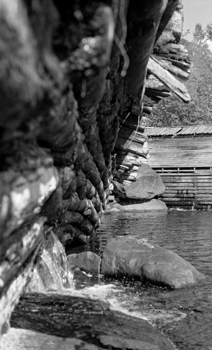 Framveggen på Syversætredammen i elva Flisa i Åsnes i Solør, fotografert fra nordvestre elvebredd i 1955.  Fotografiet er tatt i en tørkeperiode midt på sommeren, da vannføringa i elva var minimal, slik at damkonstruksjonen var godt synlig, uten overløpende vann.  Dermed ble forfallet og råteskadene på tømmerkonstruksjonen tydelige.  Den var åpenbart var moden for fornyelse.  Syversætredammen var en terskeldam som var bygd øverst i fossen, åpenbart for å skape et vannreservoar for det nedenforliggende kvernbruket, muligens også for å heve vannspeilet og lette tømmerfløtinga i det ovenforliggende elveløpet.  Dammen skapte et fall på cirka to meter.  SJF.1989-02316 viser den samme dammen mens den ennå var i god stand og på et tidspunkt da det var mye vann i Flisa.