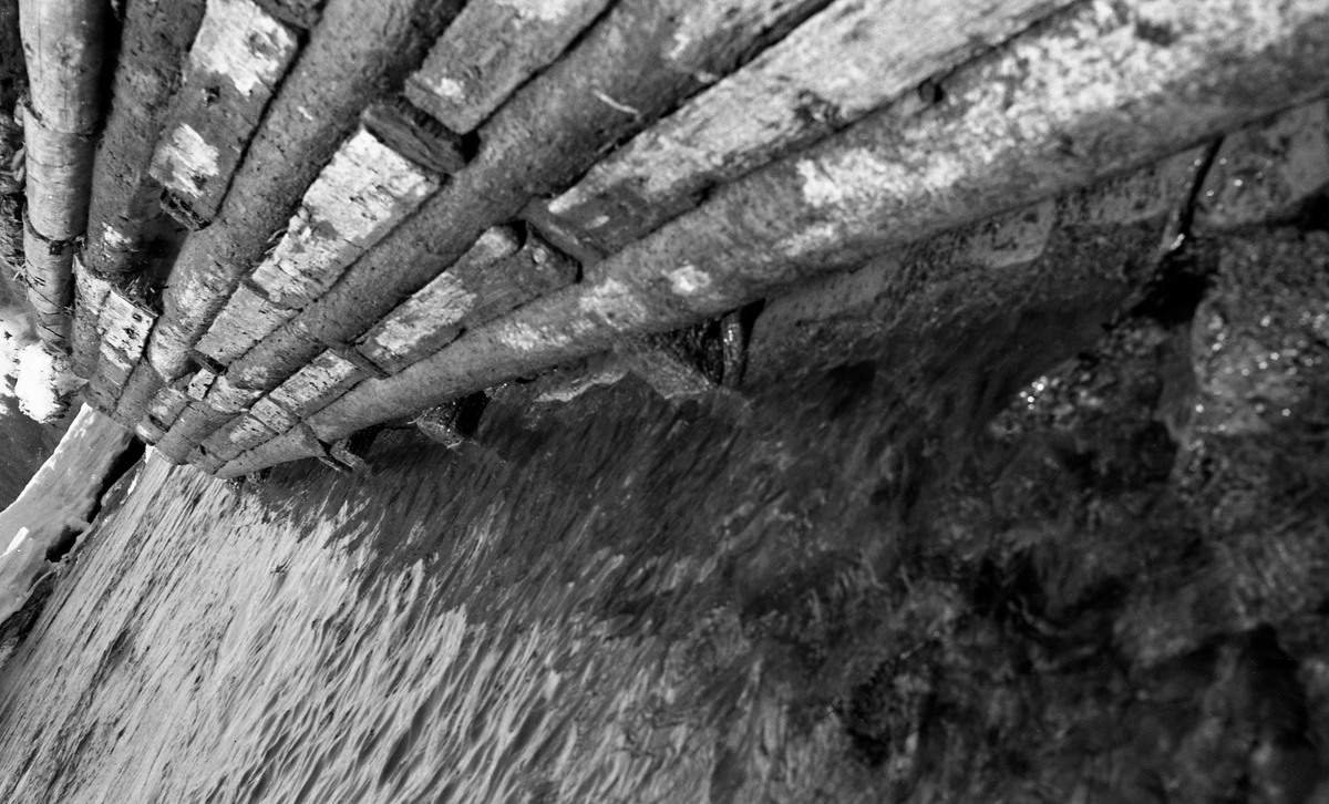 Detalj fra skådam, oppført i steinfylt tømmerkistekonstruksjon, i Bingsfossen i Sørum kommune i Akershus.  Fotografiet ble tatt i 1963, tidlig på våren mens vannstanden var lav.  I bakgrunnen lå det fortsatt digre is- og snøflak, og vannstanden Glomma var lav.  Skådammen - en ledeskjerm for fløtingstømmeret - var laftet som ei rektangulær kiste på berggrunnen i elvefaret.  Den hadde en del tverrstokker, som, når konstruksjonen ble fylt med stein fra elvefaret, bidro til at de sto støtt til tross for press fra flomvann og tømmer.  Stokkene i tømmerkista var felt sammen med luftige mellomrom, som her var forsøkt tettet med bordstubber, antakelig for å forebygge at tømmerstokker skulle hekte seg fast i veggen på skådammen.