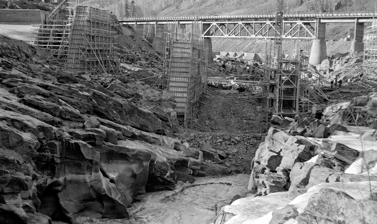 Anleggsarbeid i forbindelse med bygginga av et kraftverk ved Harpefossen i Gudbrandsdalslågen, i Sør-Fron kommune i Oppland fylke, i 1964.  Fotografiet er tatt mot et nesten tørrlagt elveleie i et trangt parti med bratte, opprevne bergskrenter på begge sider av elveløpet.  Da dette fotografiet ble tatt var man i ferd med å forskale og støpe betongfundamenter, som antakelig skulle inngå i en damkonstruksjon.  I bakgrunnen (øverst i bildeflata) ser vi ei stålbru på betongfundamenter, med fagverksunderstøtting under det langt midtspennet.  Under dette spennet skimter vi en skådam av armert betong som ble støpt for å lede hovedstrømmen i elva mot inntaktspunktet for kraftverkets turbiner.  Kraftstasjonen ved Harpefossen har to kaplanturbiner og en årsproduksjon på 427 GWh.  Anlegget utnytter et fall på 34 meter i Lågen.  Det ble satt i drift i 1965.