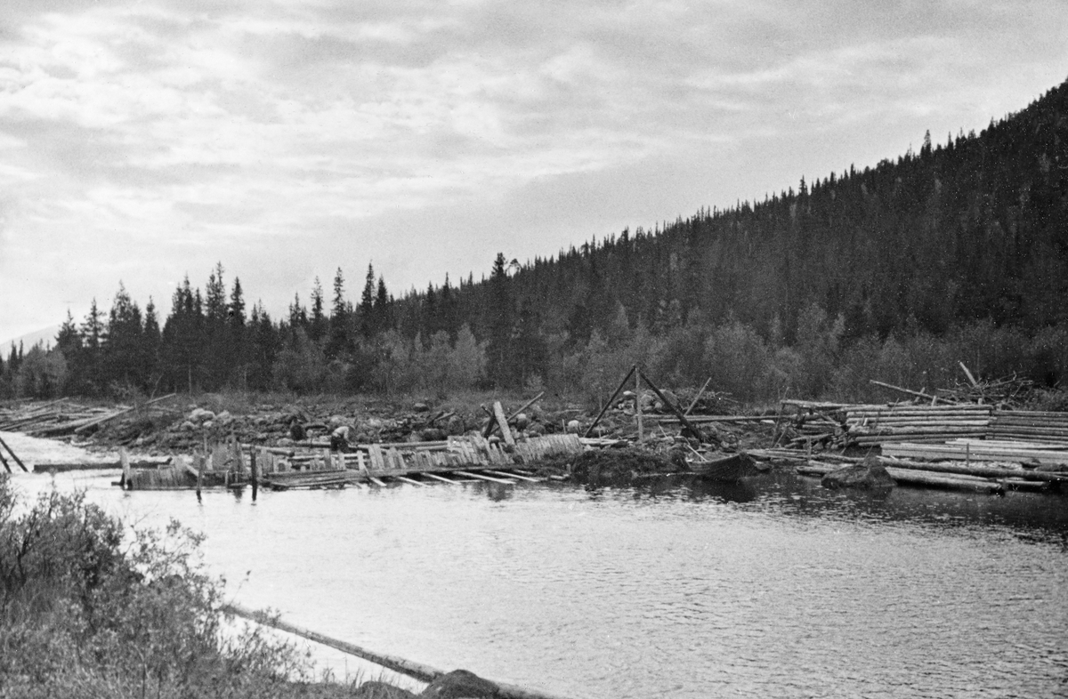 Reparasjon av Finnstaddammen i Finnstadåa (øvre del av Unsetåa) i Øvre Rendalen i Hedmark.  Fotografiet er tatt i medstrøms retning fra en posisjon på østsida av vassdraget en dag i slutten av september 1934.  I forgrunnen ser vi vannspeilet på dammen, som var ganske nedtappet på opptakstidspunktet.  På damstedet stikker en del plank opp, som en nedre avgrensning for det stillefløytende vannet.  Dette er muligens rester av en eldre damkonstruksjon.  Til høyre for denne konstruksjonen ser vi et trebeint stativ, antakelig en stubbebryter med talje, som gjorde det overkommelig å løfte tunge stokker og digre steiner.  Bakenfor skimtes en mannsskikkelse i arbeid. Til høyre for eksisterende dam (på vestsida av elva) later det til å være oppført ei tømmerkiste til en ny dam.