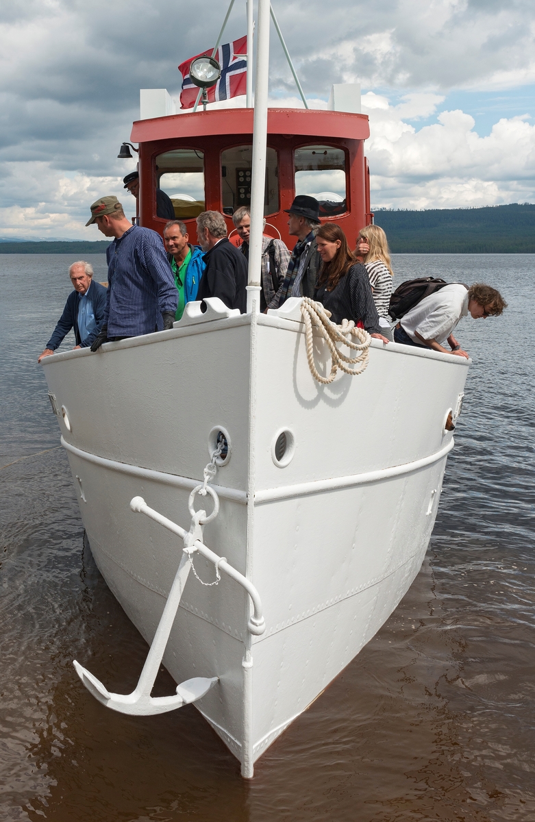 Slepebåten «Trysilknut» fotografert på den 47 kvadratkilometer store Osensjøen i grensetraktene mellom Åmot og Trysil i Hedmark, der Trysilknut gikk som tømmersleper fra 1914 til 1984.  Etter den tid har den 55 fot lange og 12 fot brede båten vært brukt som museumsbåt.  Den ble restaurert i perioden 2011-2014, og dette fotografiet er fra den første turen båten gjorde med passasjerer etter at restaureringa var avsluttet med maling av eksteriøret.  Skroget er kvitmalt og overbygningen (styrhus og bysse) er rødbrun.  Disse fargene har Trysilknut hatt siden båten ble ombygd fra damp- til dieseldrift i 1957-58, bortsett fra at vulsten som markerer hvor rekka møter den nedre delen av skroget lenge var grønnmalt.  Fotografiet er tatt mot båtens baugparti idet den legger til ved brygga på Sørlistøa fløtermuseum etter en rundtur på sjøen som inngikk i markeringa av Trysilknuts 100-årsjubileum 11. juni 2014.  På fordekket ser vi blant annet pensjonert museumsdirektør Tore Fossum (som ledet Norsk Skogbruksmuseum da Sørlistøa og Trysilknut ble innlemmet i museet), maskinist og omviser Allan Tutvedt, driftsleder Kjell Magne Kaveldiget fra Norsk Skogmuseum, direktør Harld Jacobsen fra Anno museum AS, konservator Bjørn Bækkelund (bak flaggstanga), avdelingsdirektør Stig Hoseth og fagkonsulent Ingvild Herberg fra Norsk Skogmuseum, regnskapsmedarbeider Ylva Røinaas fra Anno museum og seniorrådgiver Erik Småland fra Riksantikvaren.  Mer om Trysilknut og dens historie under fanen «Andre opplysninger».