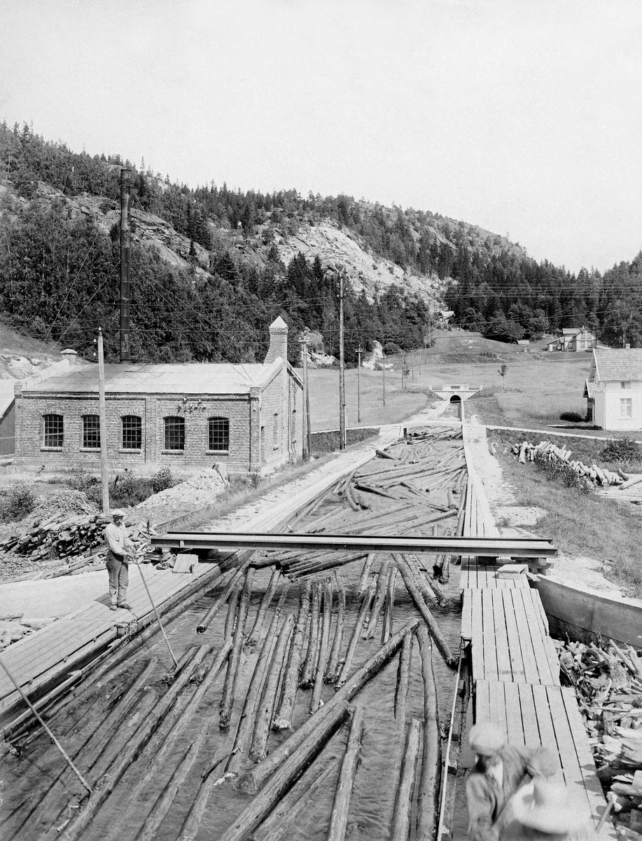Fra utløpet av den drøyt tre kilometer lange tømmertunnelen i Glommas vestre løp, mellom Isnesfjorden og Visterflo.  Fotografiet er tatt fra en posisjon over den åpne kanalen fra tunnelens utløp til lenseanlegget i Visterflos strandsone.  Vi ser hvordan tømmeret kommer flytende løst fra den ovenforliggende delen av vassdraget mot et anlegg der stokkene skulle «moses» (buntes) for videre sleping nedover vassdraget.  Bildet viser hvordan fløterne gikk på bordganger langs den åpne kanalen med langskaftete fløterhaker, som ble brukt til å «rette» tømmeret slik at stokkene fløt noenlunde parallelt mot det nedenforliggende moseanlegget.  På tvers av kanalløpet lå det smale, rekkverksløse bruer som gjorde det mulig for fløterne å krysse uten å gå spesielt langt.  Den teglmurte bygningen tmed jerninnfattete vinduer og høyt, bardunert skorsteinsrør, gav ly for dampmaskinen som drev mosemaskinene.  Ved høyre bildekant skimter vi enden av kontorbygningen ved Eidet lense.  I bakgrunnen til høyre ser vi våningshuset på småbruket som avgav grunn til utløpskanalen.
