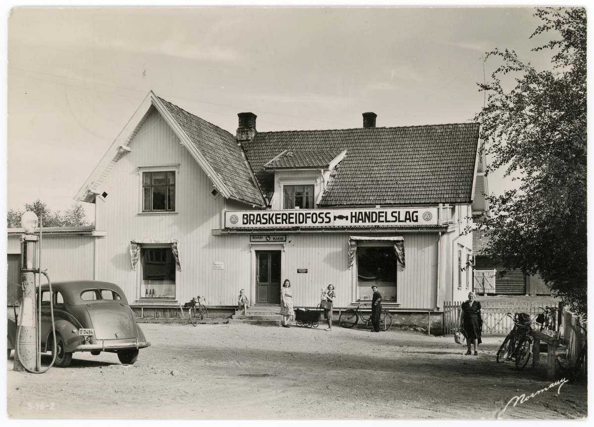 Forretningsgården til Braskereidfoss handelslag i Våler i Hedmark.  Det dreier seg om en bordkledd bygning, muligens en reisverkskonstruksjon, som lå litt tilbaketrukket fra Braskereidvegen med en grusplass foran.  På denne plassen, til venstre i bildet, var det ei bensinpumpe, der det sto en bil. Dette er en Ford V8 fra 1937, antakelig fotograf Normanns eget kjøretøy. Forrtningsgården ser opprinnelig ut til å ha vært oppført i vinkel. i halvannen etasjes høyde, med forholdsvis bratte saltak som var tekt med falset teglstein.  Da dette fotografiet ble tatt var det imidlertid oppført et enetasjes tilbygg med pulttak i vinkelen mellom de to opprinnelige bygningsfløyene, sannsynligvis for å kunne utvide ekspedisjonslokalene.  På taket av tilbygget var det montert et langt. kvitt skilt der forretningens navn sto skrevet med mørke bokstaver, supplert med symboler som viste en kjedetilknytning til NKL (Norges Kooperative Landsforening - samvirkelagskjeden).  Også til venstre på bildet ser vi et lavt, bølgeblikktekket tilbygg.  Bygningen hadde tradisjonelle krysspostvinduer i andre etasje, men noe større, sprosseløse «utstillingsvinduer» på den fasaden som vendte mot Braskereidvegen.  Da dette fotografiet ble tatt var det folk med sykler og ei barnevogn på plassen framfor forretningen.  Til høyre bak forretningsbygget ser vi et laftehus som tjente som uthusbygning for Handelslaget.  

Da dette fotografiet ble tatt var Handelslaget den eneste kolonialbutikken i tettstedet Braskereidfoss.  Kari Gravdal, som drev blomsterforretning, hadde riktignok også noen varer i tillegg til det som var hennes hovedsortiment.  Ellers var det forretninger i Nordhagen (Karlstad-butikken), på Gisti (Birger Elsetrønning) og i Tørråsen (Strandli handel).

Forretningslokalene til Braskereidfoss handelslag lå ved det som seinere er blitt hovedinnkjørselen til trelastbedriften Våler skurlag (Moelven Våler).