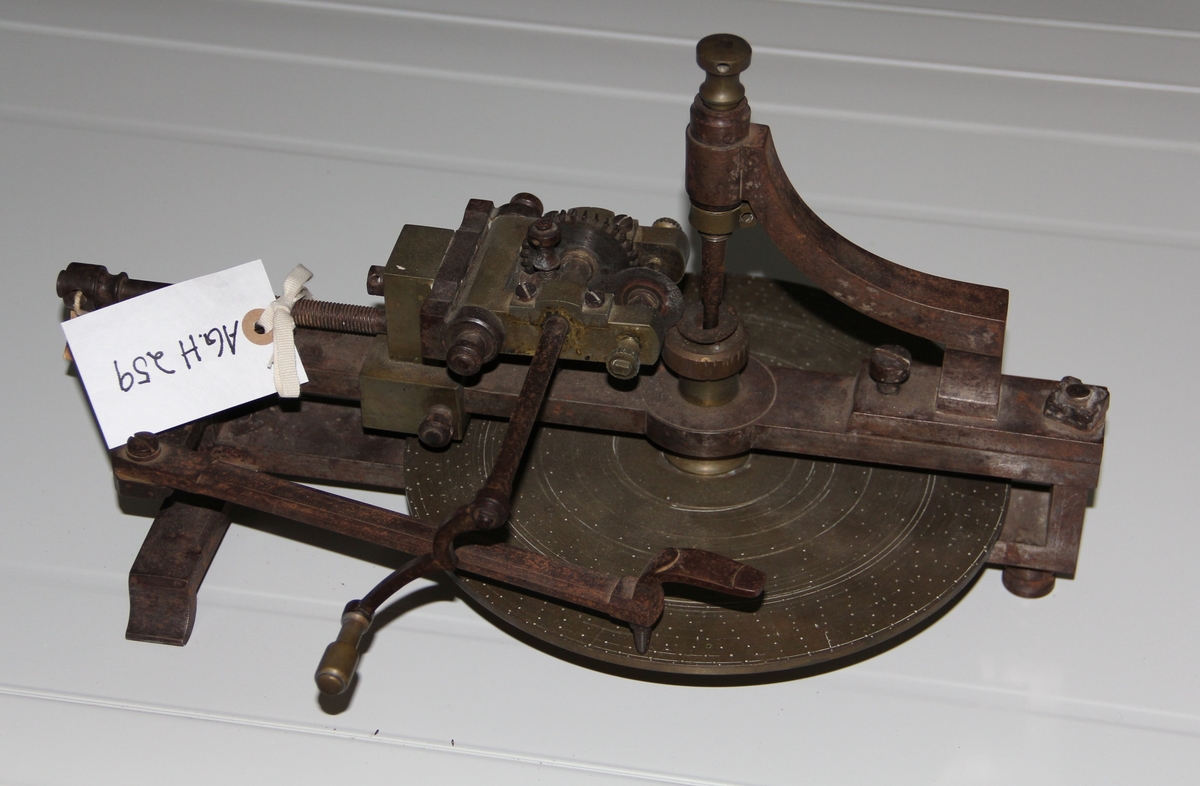 Messing og jern skive, brukt til å frese ut tagghjul til gulvklokke. Ser ut som en sirkulær skive inni en komplisert hånddrevet maskin.