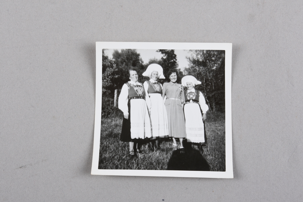 Sorthvitt fotografi av fire kvinner utendørs på gressplen med trær i bakgrunnen. Tre er iført Fanabunad, to av dem med hodeplagg. Den fjerde er iført pen kjole og lyse sko. Skyggen av fotografen vises i forgrunnen til venstre.