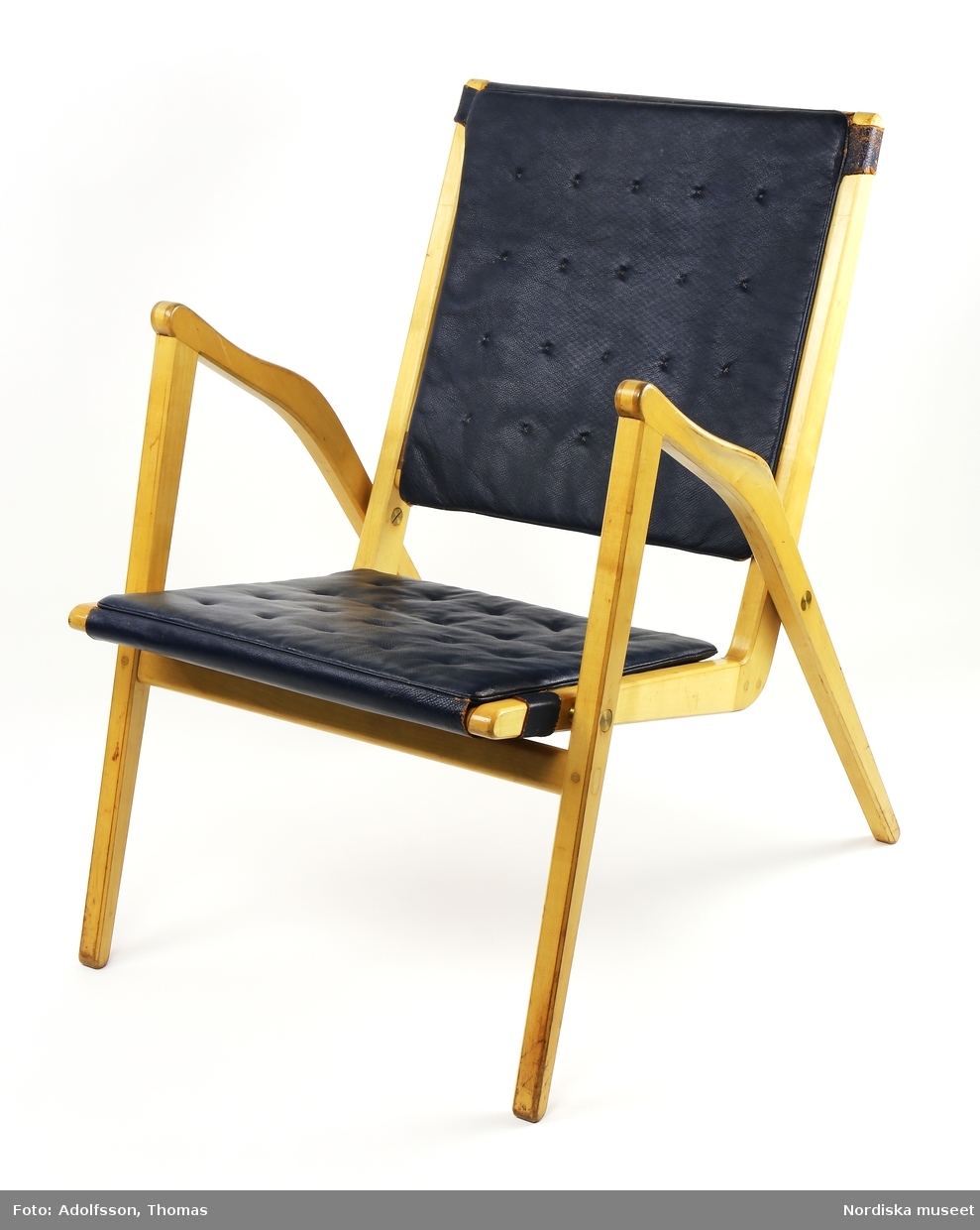 Stapelbar stol, karmstol, med stativ av klarlackad björk. Formpressad, perforerad sits och rygg av valnötsfaner. Sittdel och stativ sammansatta med mässingsbeslag av tryckknappstyp. Sammanfogning med slitsar som även ger en dekorativ verkan. Formgiven 1955 och sannolikt tillverkad samma år.
/Anna Arfvidsson Womack 2019-01-29