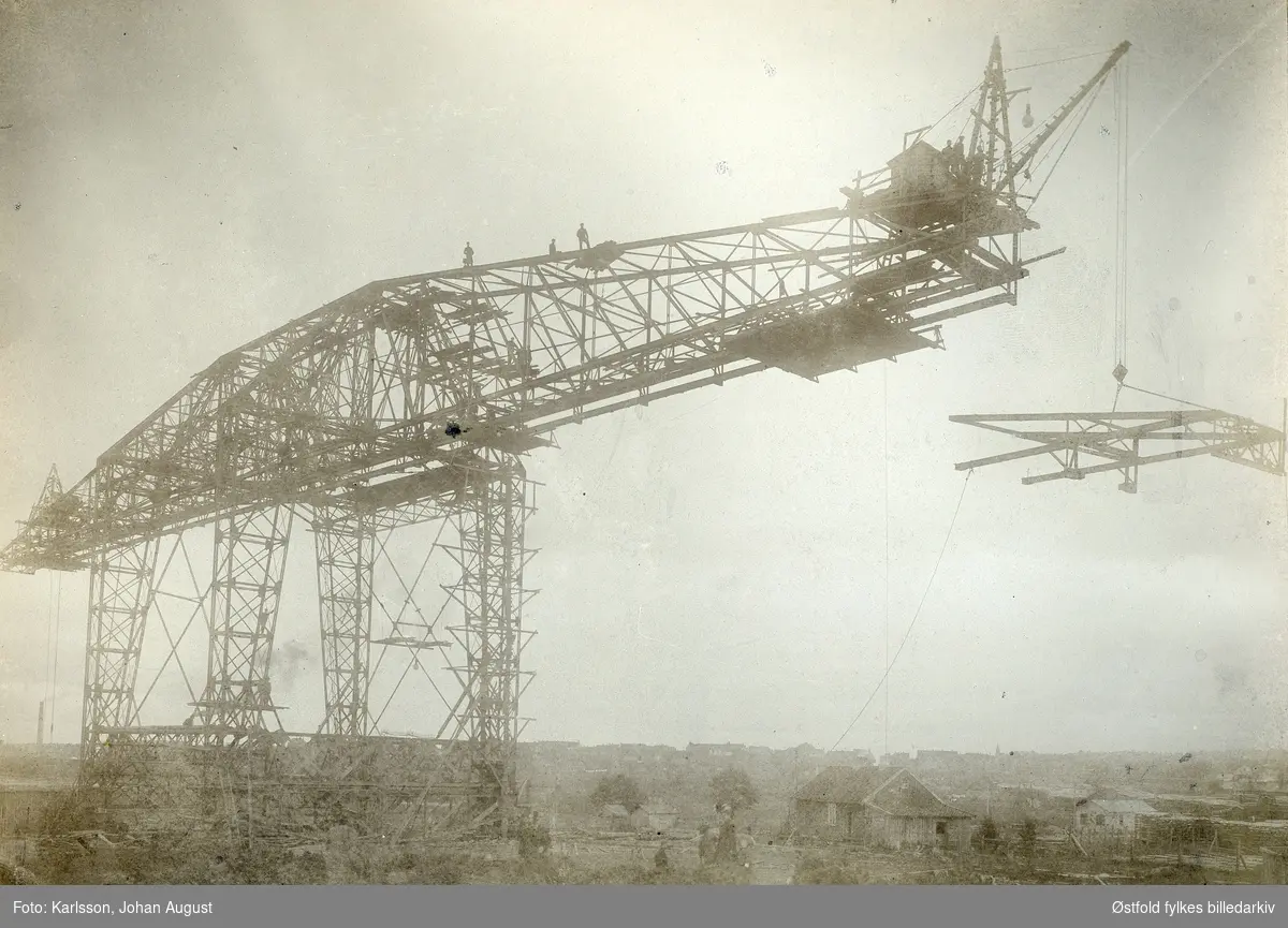 Tømmerkrana og tømmeropplag på Opsund i  Sarpsborg 1900. Luftbane til høyre.
Flere menn står på toppen av krana og ved kranhuset.