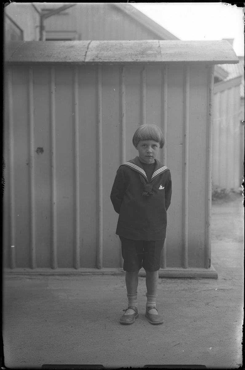 En pojke i sjömansdräkt står framför ett träplank eller en träbyggnad. I fotografens egna anteckningar står det "[Beställning genom] Axel, pojken".