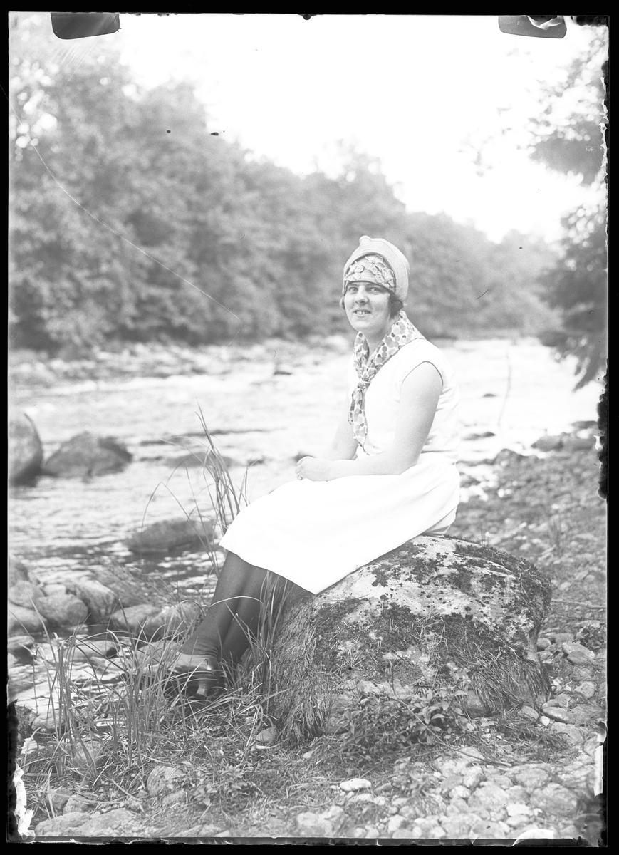 Lizzie Skoglund sitter på en sten vid vattenbrynet och ler mot kameran. Hon har på sig en vit ärmlös klänning, ljus hatt, mönstrad sjal och svarta strumpor och skor.