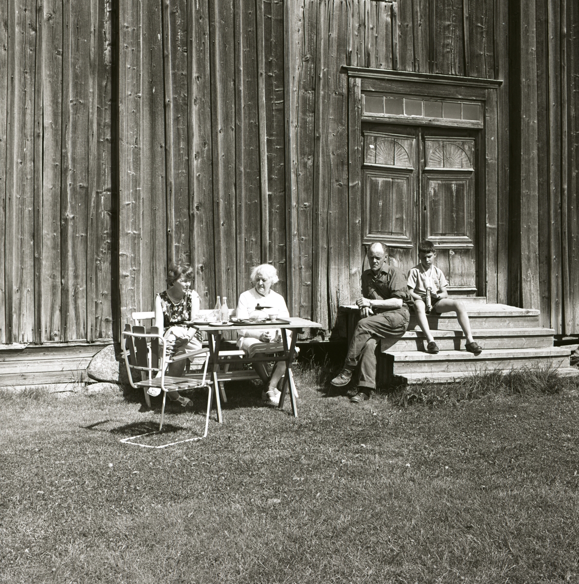 Två kvinnor sitter vid ett bord framför en byggnad och till höger om dem sitter en pojke och en man på en trappa, 1967-68.