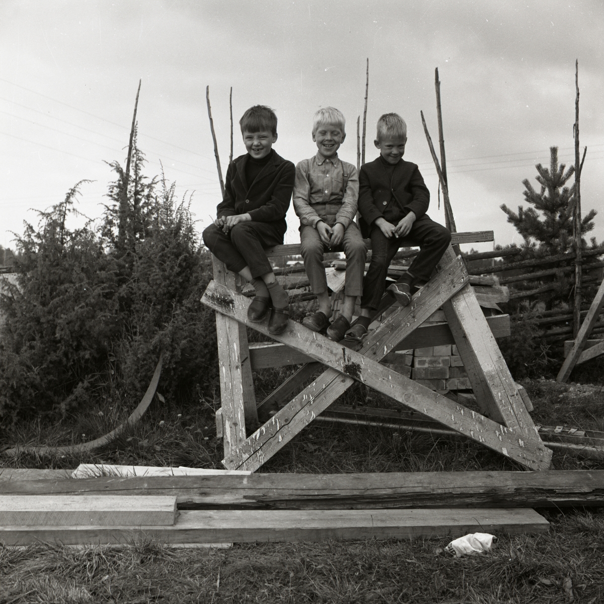Tre pojkar sitter på en träbock och ler vid gården Sunnanåker, 1967-68.