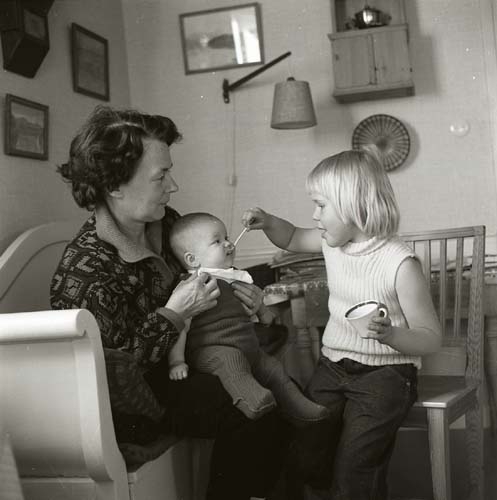 En kvinna sitter i en soffa med ett barn i sin famn. Framför dem sitter ett äldre barn och matar det yngsta barnet med en sked, 1970.