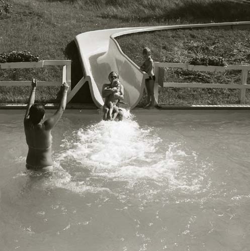 Ett barn och en vuxen person åker i en vattenrutschkana ned i en vattensamling. I vattnet står en annan person och väntar med armarna resta mot himlen, 1983.