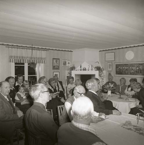 En grupp människor har samlats i ett rum och sitter intill väggarna samt vid bord, 1984.