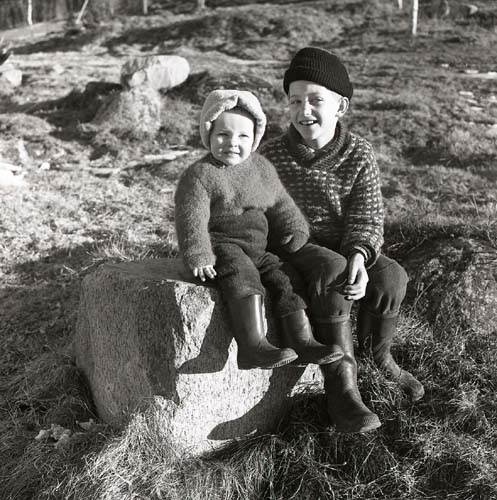 Två barn i olika åldrar sitter på en sten. Det äldre barnet bär en stickad tröja, 1961.