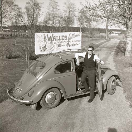 En man poserar framför en bil och en reklamskylt. Bilen är placerad tvärs över en väg, 1964.