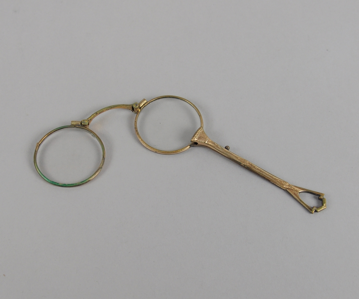 To brilleglass med metallinnfatning og metallhåndtak. Brilleglassene kan brettes sammen, og en liten knapp på håndtaket som kan trykkes inn for å skille de to brilleglassene.