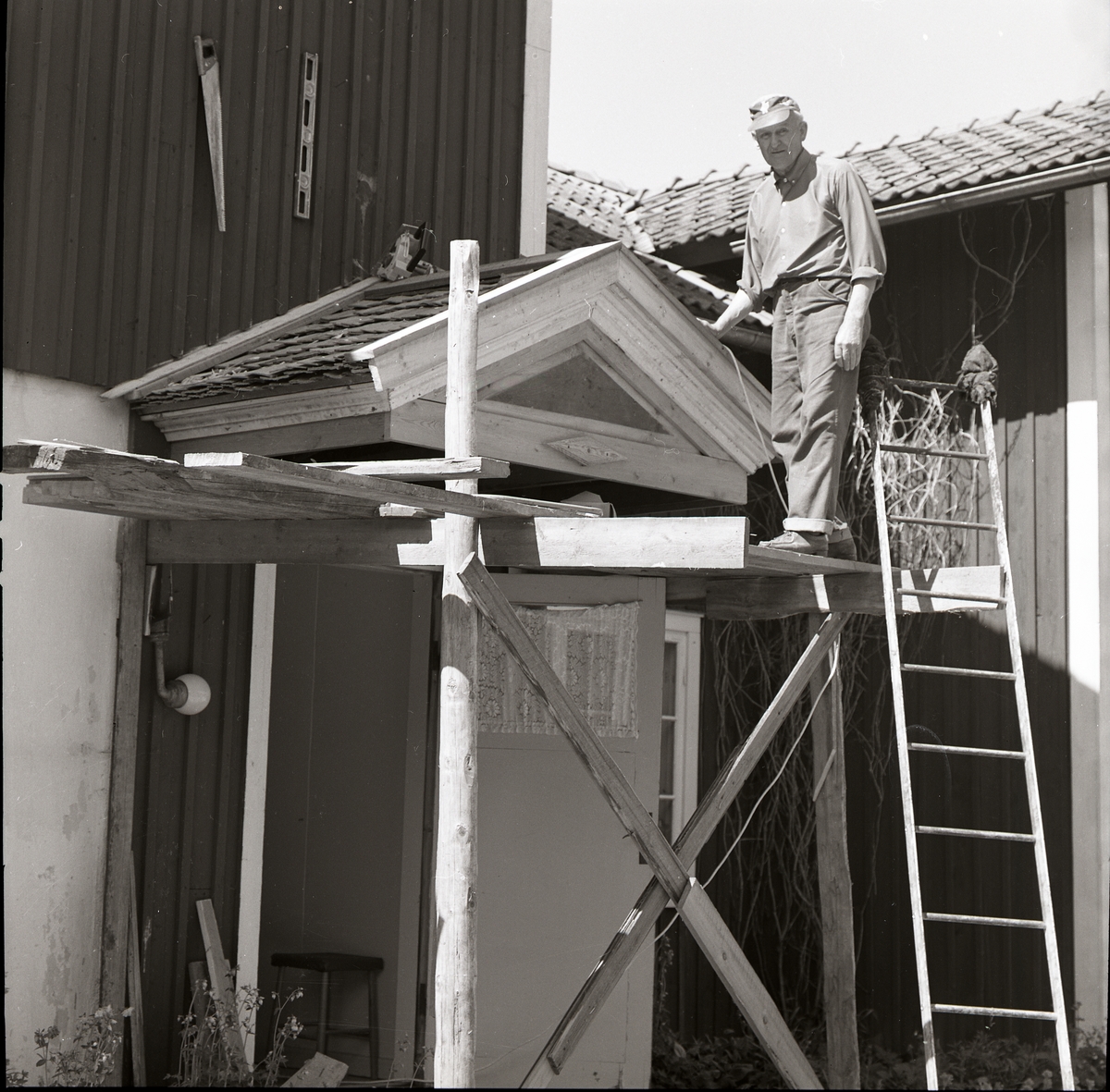 En man med keps står på en byggnadsställning och reparerar ett litet sadeltak. Bredvid honom står en stege lutad mot ställningen. På väggen hänger diverse verktyg, Sunnanåker 12 juni 1979.