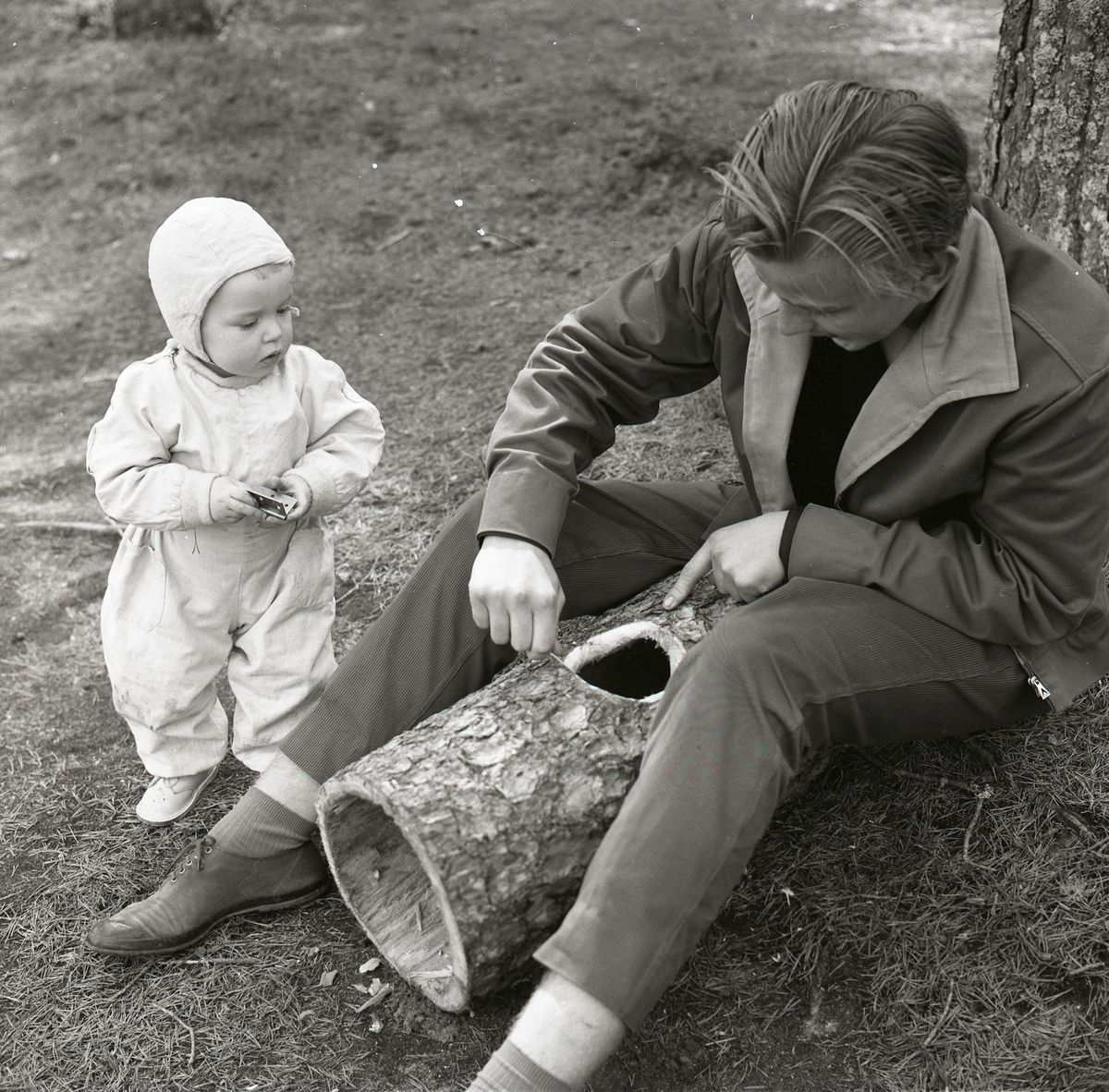 En pojke sitter på marken intill en trädstam och täljer till en rund öppning i en holk med en kniv. Ett barn står bredvid hållandes ett gångjärn och tittar intresserat på pojkens aktiviteter, 10 maj 1959.