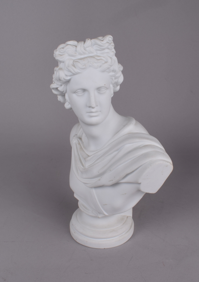Klassisk mannsfigur med håret i sløyfe og draperier rundt skuldrene. Den romerske/greske guden Apollo