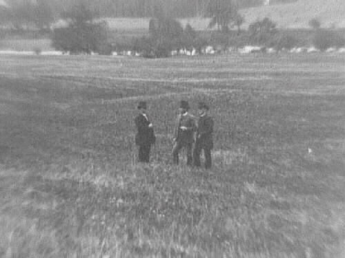 Alfshög socken i Vessigebro. Tre män står på ett fält med ån och kyrkåsen i bakgrunden.