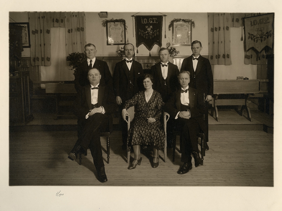 Gruppbild av 6 män och 1 kvinna, kvinnan är sittande på en stol i mitten, i I.O.G.T:s lokaler. Bakom dom syns 3 flaggor med texten I.O.G.T samt 2 texttavlor. Sittande t.v: Ludvig Andersson (1887-1984). 
Enligt påskrift "IOGT-lokalen d 7/2 1932".